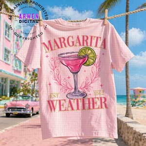 May include: A light pink t-shirt with a margarita design. The design includes a pink cocktail in a stemmed glass with a lime slice, surrounded by a floral wreath. The text "MARGARITA WEATHER" is printed on the shirt.