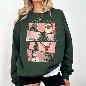 May include: Dark green sweatshirt featuring a festive Christmas design. The graphic includes Santa Claus, a decorated Christmas tree, a snowman, and a reindeer, all set against a pink backdrop. The sweatshirt has a white undershirt peeking out from the bottom.