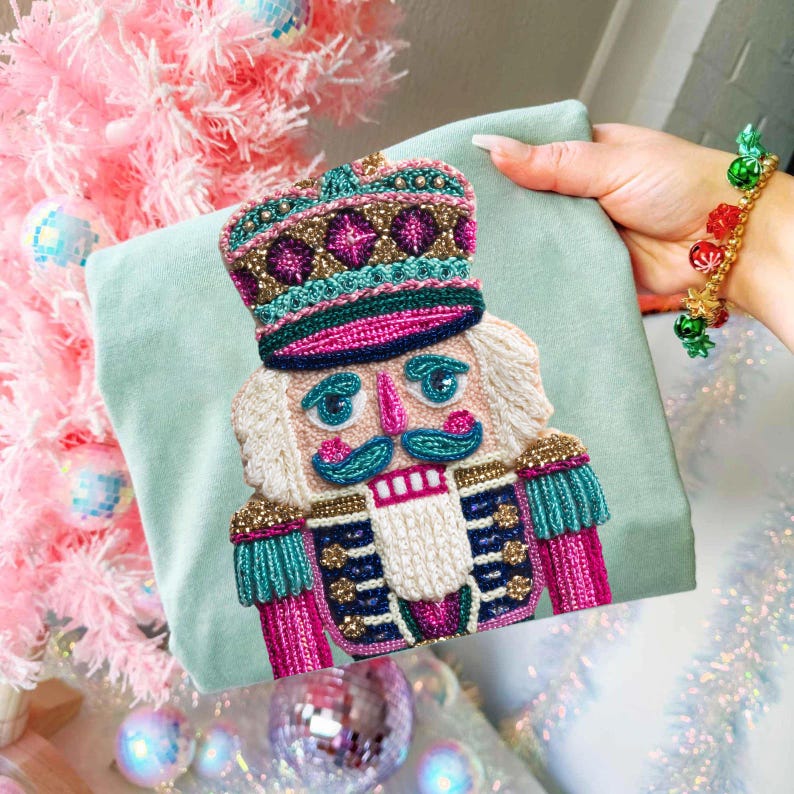 May include: A light green t-shirt featuring a detailed, embroidered nutcracker design. The nutcracker has a colorful hat, uniform, and mustache. The design incorporates pink, teal, gold, and blue accents. The shirt is held up in front of a pink Christmas tree.