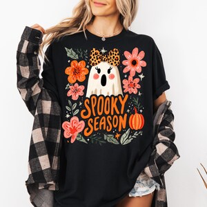 K&ouml;nnte beinhalten: Schwarzes T-Shirt mit einem Halloween-Motiv. Das Design zeigt einen wei&szlig;en Geist mit Leopardenmuster-Schleife, orangefarbenen und rosa Blumen, einen K&uuml;rbis und die Worte "Spooky Season".