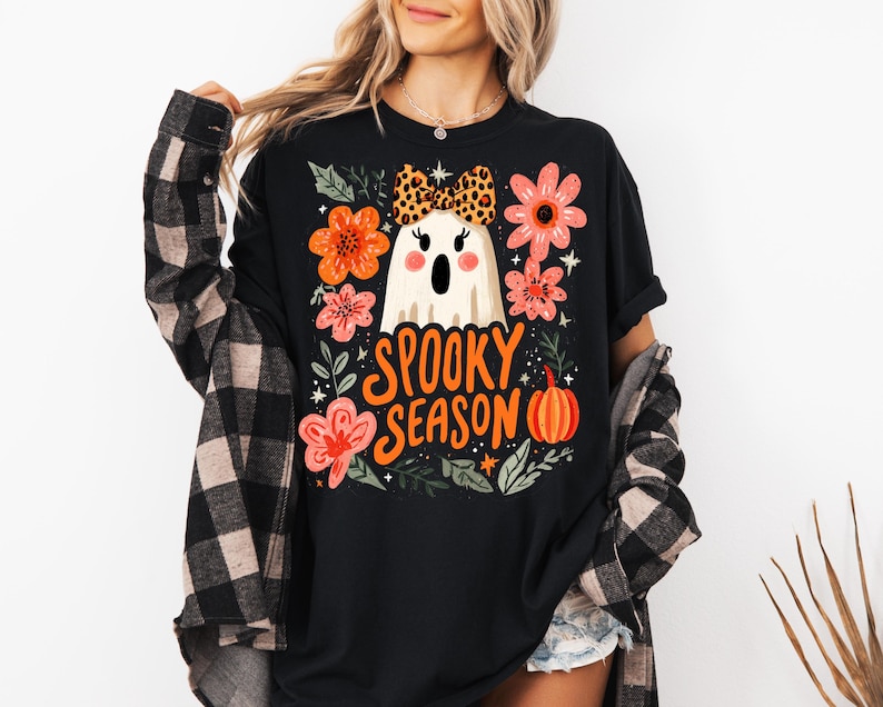 K&ouml;nnte beinhalten: Schwarzes T-Shirt mit einem Halloween-Motiv. Das Design zeigt einen wei&szlig;en Geist mit Leopardenmuster-Schleife, orangefarbenen und rosa Blumen, einen K&uuml;rbis und die Worte "Spooky Season".
