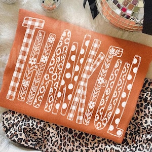 May include: A burnt orange sweatshirt featuring the word THANKFUL printed in white. Each letter displays a unique pattern, including buffalo plaid, floral vines, leopard spots, and polka dots, set against a leopard print fabric base.