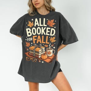 May include: Dark gray oversized t-shirt with the text "ALL BOOKED FOR FALL" in white and orange. The graphic includes fall leaves, books, a pumpkin, a mug, and glasses. A cozy autumn-themed design.