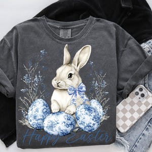 May include: Dark gray t-shirt featuring a white bunny with a blue bow, surrounded by blue floral accents and Easter eggs. The words "Happy Easter" are printed in blue. The shirt is paired with jeans and a phone case.
