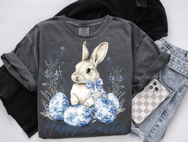May include: Dark gray t-shirt featuring a white bunny with a blue bow, surrounded by blue floral accents and Easter eggs. The words "Happy Easter" are printed in blue. The shirt is paired with jeans and a phone case.