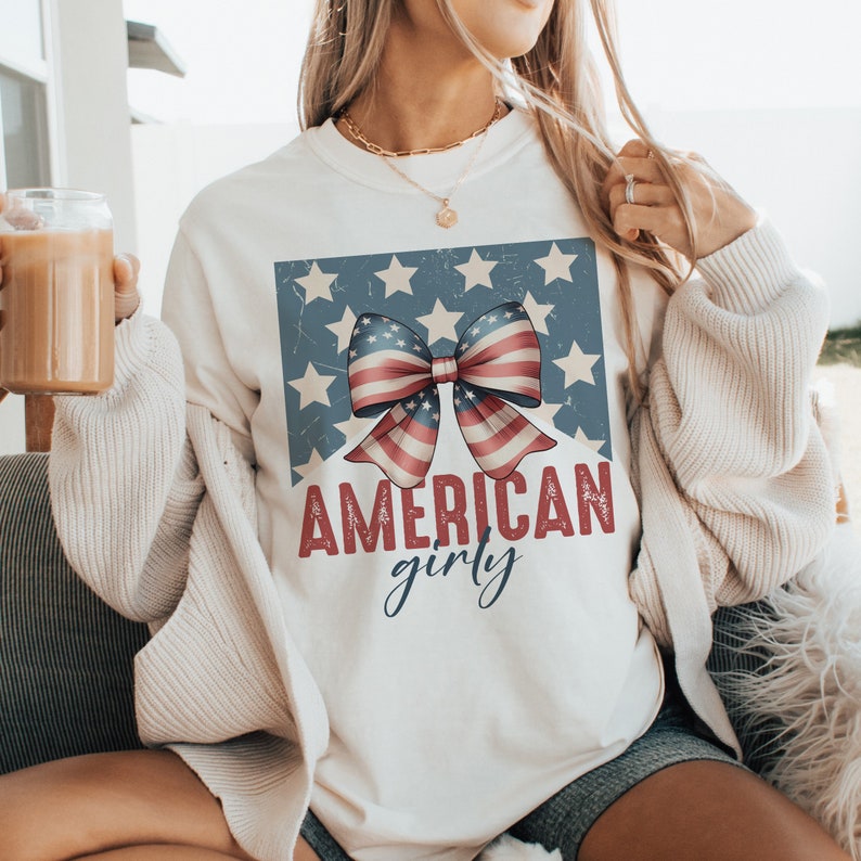 May include: White t-shirt with a red, white, and blue bow and stars design. The text "AMERICAN girly" is printed below the bow.
