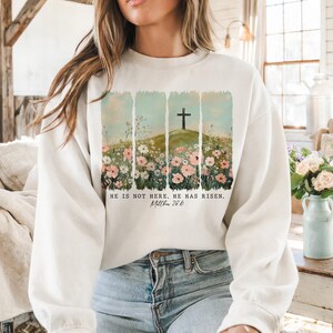 May include: White sweatshirt with a floral design and a cross on a hill. The text "He is not here. He has risen. Matthew 28:6" is printed on the sweatshirt.