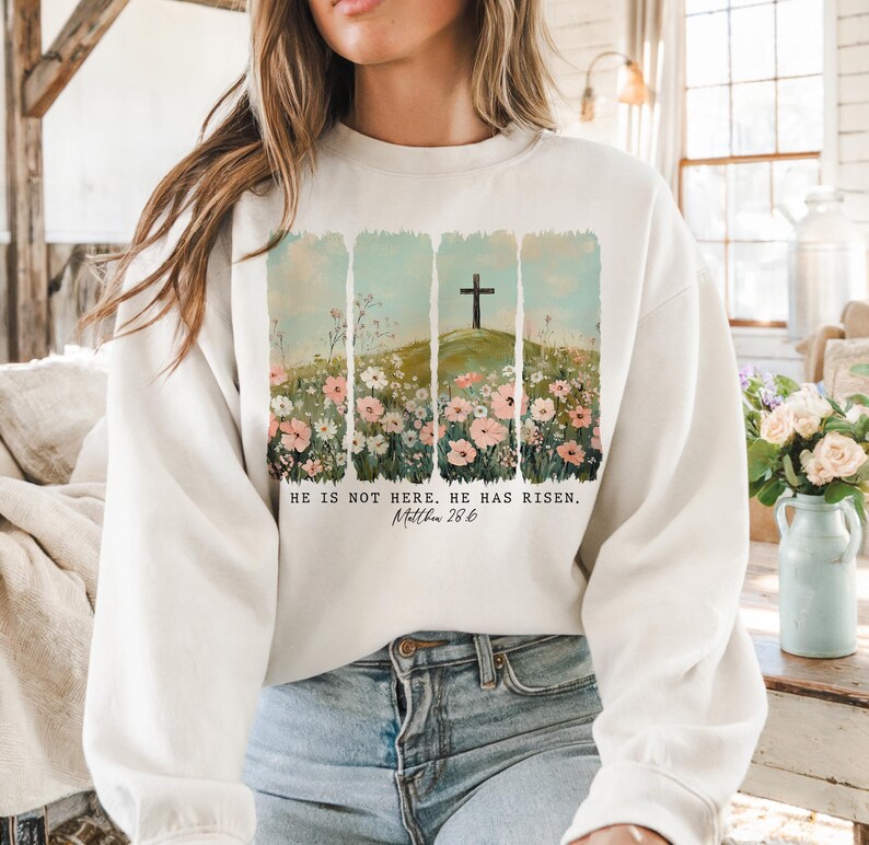 May include: White sweatshirt with a floral design and a cross on a hill. The text "He is not here. He has risen. Matthew 28:6" is printed on the sweatshirt.