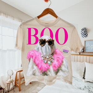 May include: A beige t-shirt featuring a graphic of a ghost wearing leopard print sunglasses, a large crystal statement necklace, and a bright pink feather boa. The word BOO is printed in large, bold, hot pink letters above the ghost figure.