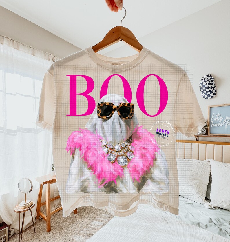 May include: A beige t-shirt featuring a graphic of a ghost wearing leopard print sunglasses, a large crystal statement necklace, and a bright pink feather boa. The word BOO is printed in large, bold, hot pink letters above the ghost figure.