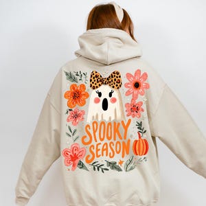 K&ouml;nnte beinhalten: Beiger Kapuzenpullover mit einem Halloween-Motiv auf dem R&uuml;cken. Das Design zeigt einen Geist mit einer Schleife im Leopardenmuster, umgeben von orangefarbenen und rosa Blumen, einem K&uuml;rbis und den Worten "Spooky Season".