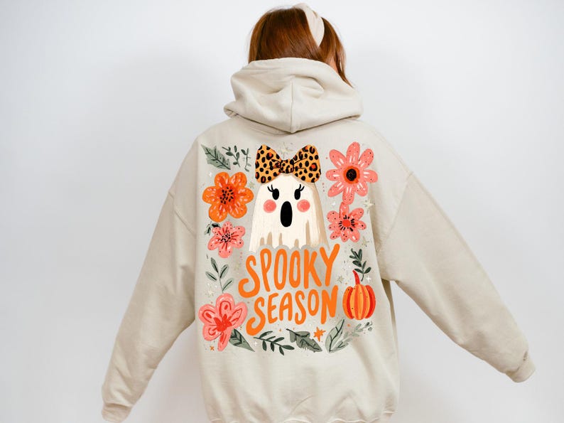 K&ouml;nnte beinhalten: Beiger Kapuzenpullover mit einem Halloween-Motiv auf dem R&uuml;cken. Das Design zeigt einen Geist mit einer Schleife im Leopardenmuster, umgeben von orangefarbenen und rosa Blumen, einem K&uuml;rbis und den Worten "Spooky Season".
