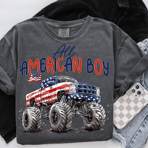 May include: Dark gray crew neck sweatshirt with the text "All AMERICAN BOY" in red and blue. The graphic features a monster truck with an American flag design. The shirt is folded with a pair of blue jeans and a phone case.