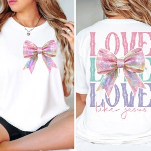 K&ouml;nnte beinhalten: Wei&szlig;es T-Shirt mit einer rosa und schillernden Schleife auf der Vorderseite und dem Text "LOVE LIKE LOVE like jesus" auf der R&uuml;ckseite.