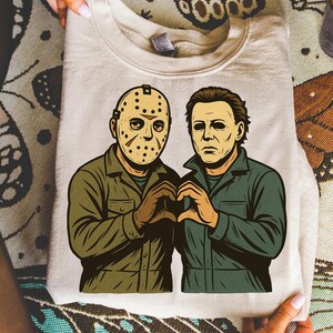 May include: A beige sweatshirt featuring a graphic of two horror movie characters, one wearing a hockey mask and the other with a blank expression. They are forming a heart shape with their hands. The characters are dressed in green and brown jumpsuits.