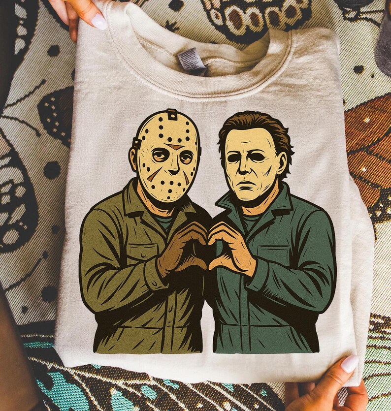 May include: A beige sweatshirt featuring a graphic of two horror movie characters, one wearing a hockey mask and the other with a blank expression. They are forming a heart shape with their hands. The characters are dressed in green and brown jumpsuits.