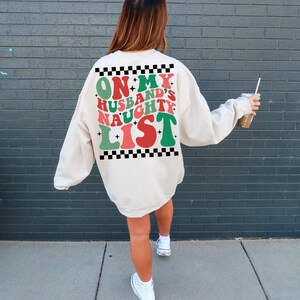 May include: Cream-colored sweatshirt with the text "ON MY HUSBAND'S NAUGHTY LIST" in red, green, and white. The text is framed by a black and white checkered pattern. A festive holiday-themed apparel item.