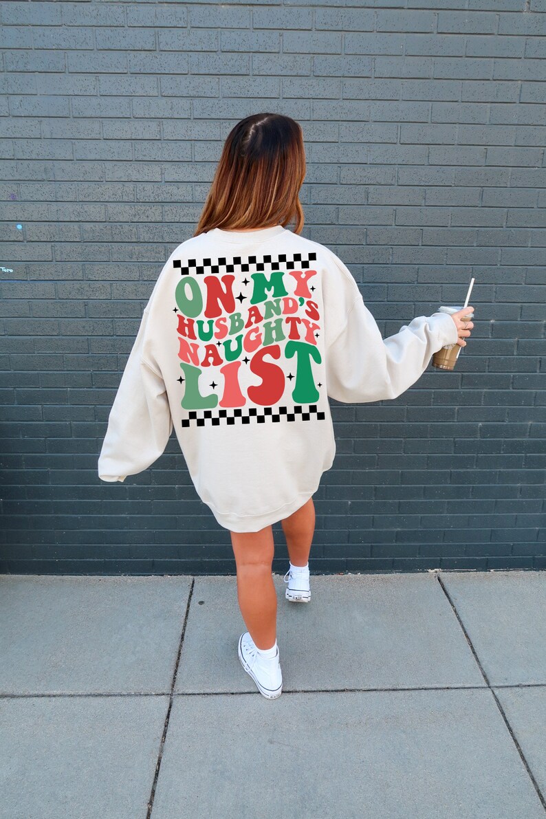 May include: Cream-colored sweatshirt with the text "ON MY HUSBAND'S NAUGHTY LIST" in red, green, and white. The text is framed by a black and white checkered pattern. A festive holiday-themed apparel item.
