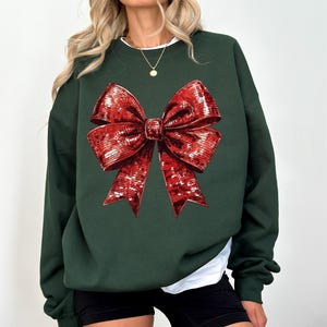 Puede incluir: Sudadera verde oscuro con un gran lazo rojo detallado. El lazo tiene una apariencia texturizada y con lentejuelas. La sudadera tiene mangas largas y cuello redondo, con una camiseta blanca debajo.