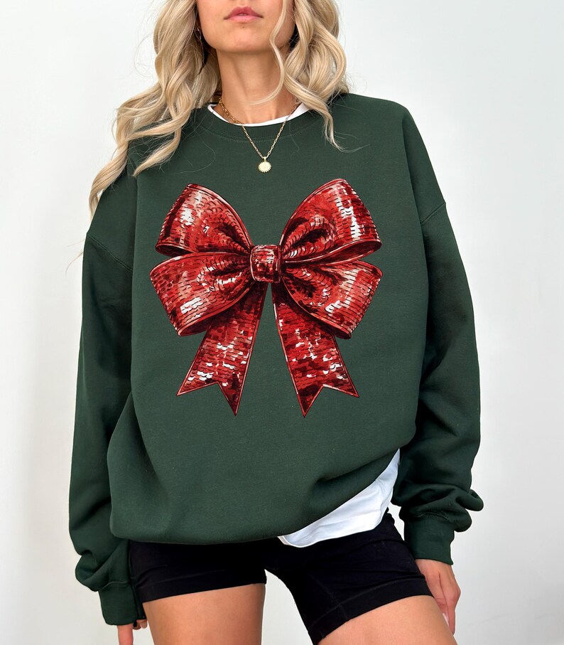 Puede incluir: Sudadera verde oscuro con un gran lazo rojo detallado. El lazo tiene una apariencia texturizada y con lentejuelas. La sudadera tiene mangas largas y cuello redondo, con una camiseta blanca debajo.