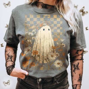 Op de afbeelding: Grijs T-shirt met een spookillustratie, bloemenkrans en bloemen. Het ontwerp staat tegen een geblokte achtergrond met een vintage esthetiek. Het shirt heeft korte mouwen.