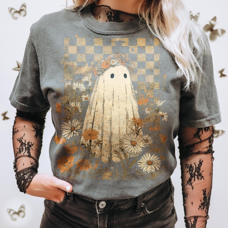 Op de afbeelding: Grijs T-shirt met een spookillustratie, bloemenkrans en bloemen. Het ontwerp staat tegen een geblokte achtergrond met een vintage esthetiek. Het shirt heeft korte mouwen.