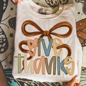 May include: Cream-colored sweatshirt with a brown bow design and the words "GIVE THANKS" in colorful, outlined letters. The text is in shades of pink, blue, and brown. The sweatshirt is folded and held by hands.