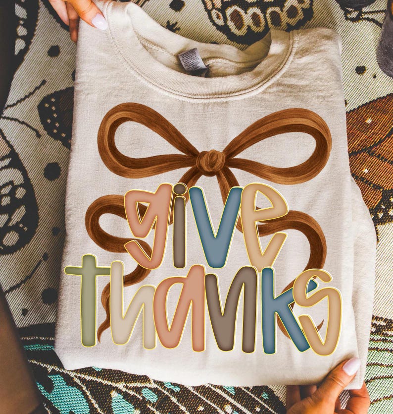 May include: Cream-colored sweatshirt with a brown bow design and the words "GIVE THANKS" in colorful, outlined letters. The text is in shades of pink, blue, and brown. The sweatshirt is folded and held by hands.