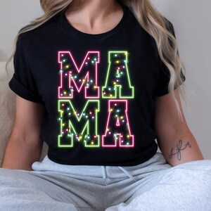 May include: Black t-shirt with the word "MAMA" in a neon-style design. The letters are outlined in pink and green, filled with small, colorful lights. The shirt is a casual style.
