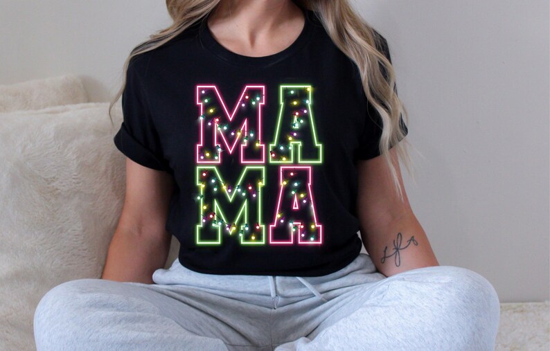 May include: Black t-shirt with the word "MAMA" in a neon-style design. The letters are outlined in pink and green, filled with small, colorful lights. The shirt is a casual style.