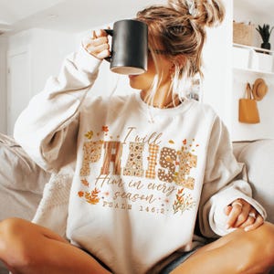 May include: A cream-coloured sweatshirt featuring the text "I will PRAISE Him in every season". The word "PRAISE" is in a patchwork style with floral and plaid patterns. The sweatshirt also has autumn-themed leaf and floral accents.