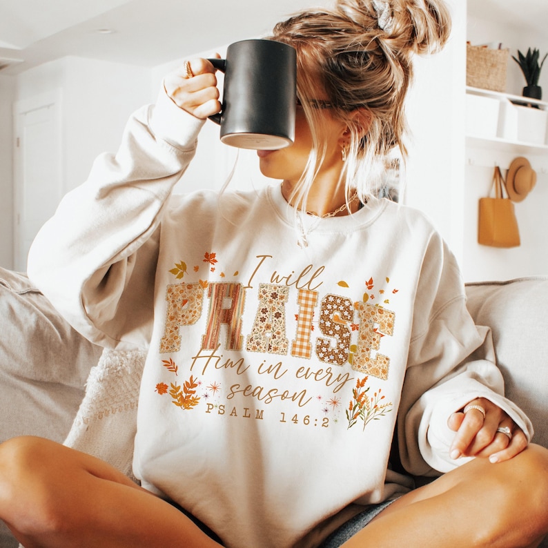 May include: A cream-coloured sweatshirt featuring the text "I will PRAISE Him in every season". The word "PRAISE" is in a patchwork style with floral and plaid patterns. The sweatshirt also has autumn-themed leaf and floral accents.