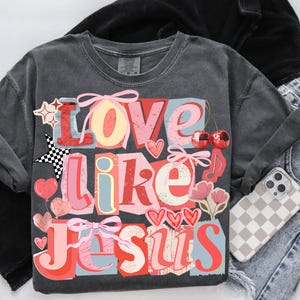May include: Charcoal grey t-shirt featuring the phrase "Love Like Jesus" in a vibrant, layered design. The graphic incorporates hearts, stars, cherries, and bows. A phone with a checkered case and denim jeans are also visible.