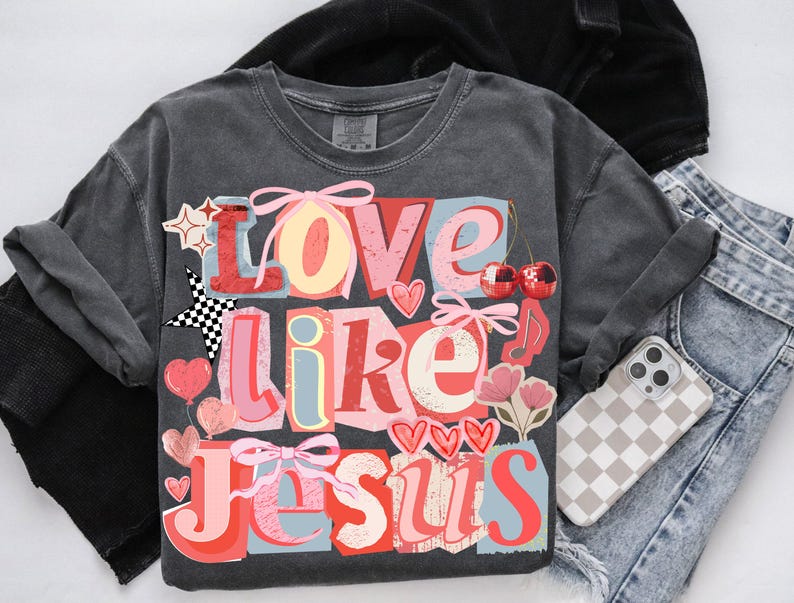 May include: Charcoal grey t-shirt featuring the phrase "Love Like Jesus" in a vibrant, layered design. The graphic incorporates hearts, stars, cherries, and bows. A phone with a checkered case and denim jeans are also visible.