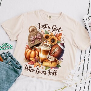 K&ouml;nnte beinhalten: Cremefarbenes T-Shirt mit einem Herbst-Themen-Grafik. Die Grafik zeigt einen K&uuml;rbis, einen Latte, einen Football und einen Kuchen, mit dem Text "Just a Girl Who Loves Fall". Eine M&uuml;tze und Sonnenblumen sind ebenfalls abgebildet.
