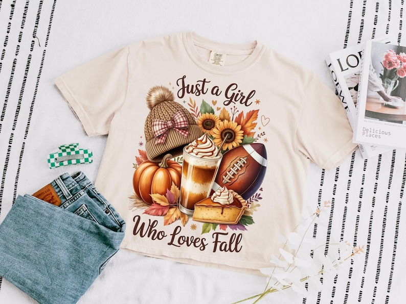 K&ouml;nnte beinhalten: Cremefarbenes T-Shirt mit einem Herbst-Themen-Grafik. Die Grafik zeigt einen K&uuml;rbis, einen Latte, einen Football und einen Kuchen, mit dem Text "Just a Girl Who Loves Fall". Eine M&uuml;tze und Sonnenblumen sind ebenfalls abgebildet.