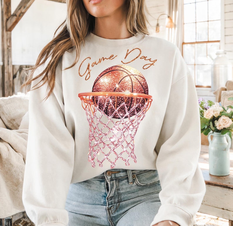 May include: White sweatshirt with the text "Game Day" in gold glitter lettering. A pink glitter basketball is going through a pink glitter basketball hoop.