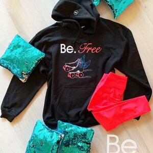 May include: Black hooded sweatshirt with the text "Be. Free" and a graphic of roller skates with wings. The sweatshirt is laid out on a wooden surface with a pair of red leggings and turquoise sequin pillows.