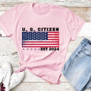 May include: Pink t-shirt with a distressed American flag graphic and the text "U.S. Citizen Est 2024".