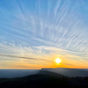 May include: A panoramic view of a mountain range at sunset. The sky is a vibrant blue with wispy clouds, and the sun is setting behind the mountains, casting a warm glow on the landscape.