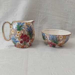 Könnte beinhalten: Ein Vintage-Set aus Sahnegießer und Zuckerdose mit floralem Muster in Creme und Gold. Der Sahnegießer hat einen Henkel und einen abgerundeten Körper. Die Zuckerdose ist rund und hat einen breiten Rand.