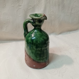 Może przedstawiać: Mały ceramiczny dzbanek w kolorze zielonym i brązowym z uchwytem. Dzbanek ma szkliwioną powierzchnię i lekko zaokrąglone ciało.