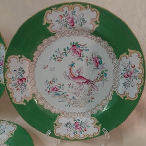 May include: A white porcelain plate with a green rim and a gold trim. The center of the plate features a colorful bird with a long tail, surrounded by flowers and leaves. The plate has a decorative border with a repeating pattern of swirls and flowers.