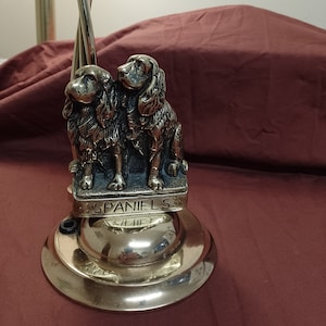 May include: A brass lamp base featuring two detailed spaniel dog sculptures. The base has a decorative design and the word "SPANIEIS" is engraved on the base. The lamp has a twisted metal pole.