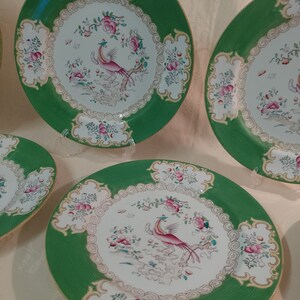 May include: Set of four green and white porcelain dinner plates with a gold rim. Each plate features a pink and white bird with a long tail, surrounded by pink and white flowers and leaves.