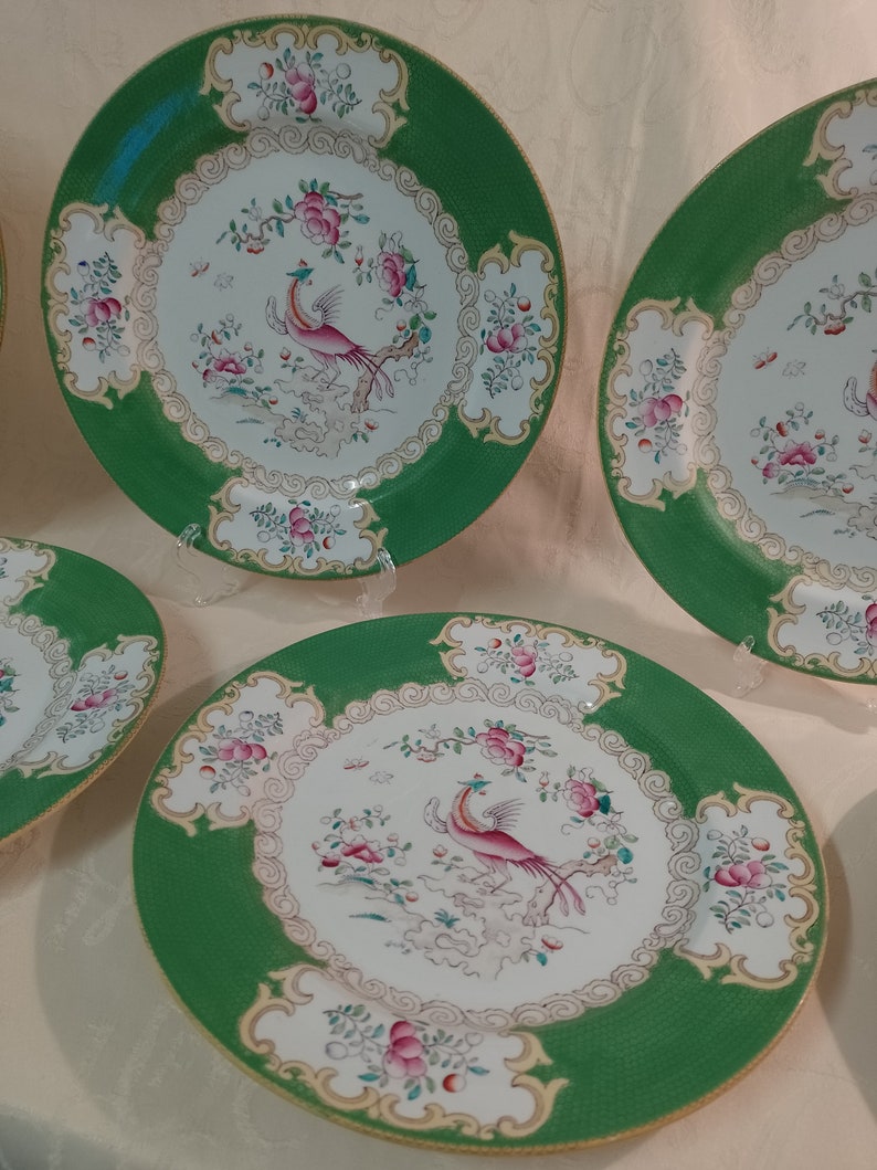 May include: Set of four green and white porcelain dinner plates with a gold rim. Each plate features a pink and white bird with a long tail, surrounded by pink and white flowers and leaves.
