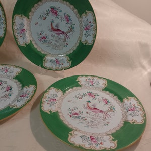 May include: Set of three green and white porcelain dinner plates with a gold trim. Each plate features a pink and white bird with a long tail, surrounded by pink and white flowers.