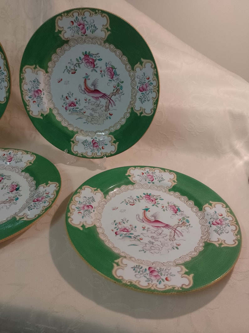 May include: Set of three green and white porcelain dinner plates with a gold trim. Each plate features a pink and white bird with a long tail, surrounded by pink and white flowers.