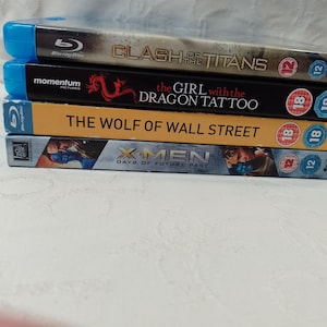 May include: A stack of four Blu-ray discs featuring the movies "Clash of the Titans", "The Girl with the Dragon Tattoo", "The Wolf of Wall Street", and "X-Men: Days of Future Past".