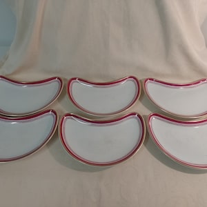May include: Six white ceramic crescent-shaped plates with a red and gold trim.